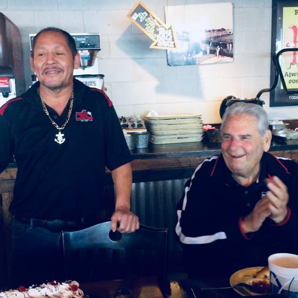 Two men, one standing and one seated, smiling in a casual restaurant setting with a cake.