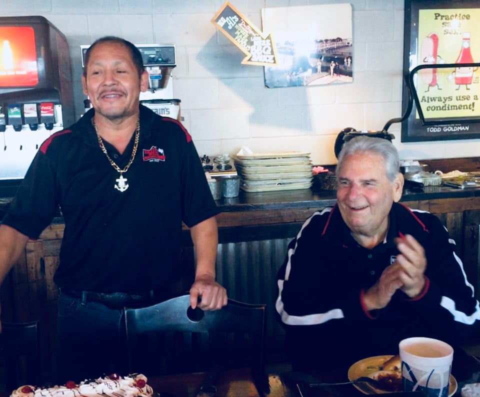 Two men, one standing and one seated, smiling in a casual restaurant setting with a cake.