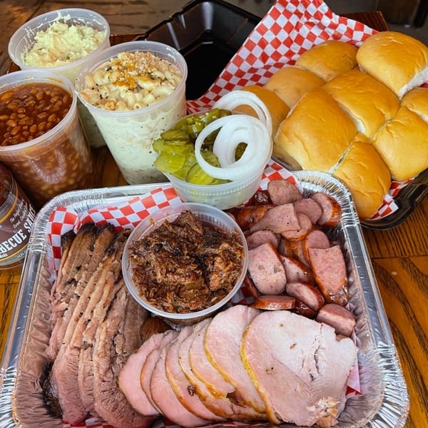 A barbecue feast with sliced brisket, ham, sausage, pulled pork, baked beans, mac and cheese, potato salad, rolls, pickles, and onions.