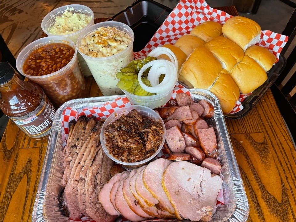 A barbecue feast with sliced brisket, ham, sausage, pulled pork, baked beans, mac and cheese, potato salad, rolls, pickles, and onions.