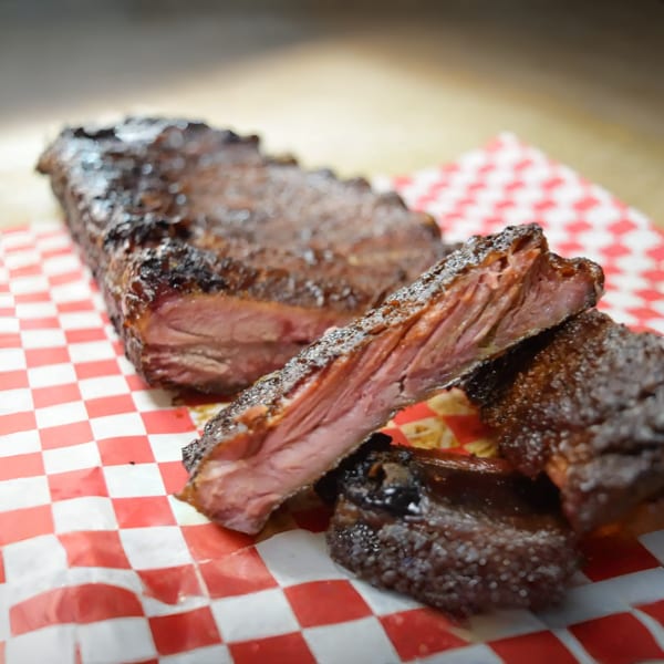Smoked ribs with a dark, crispy exterior and tender pink meat, served on red and white checkered paper.