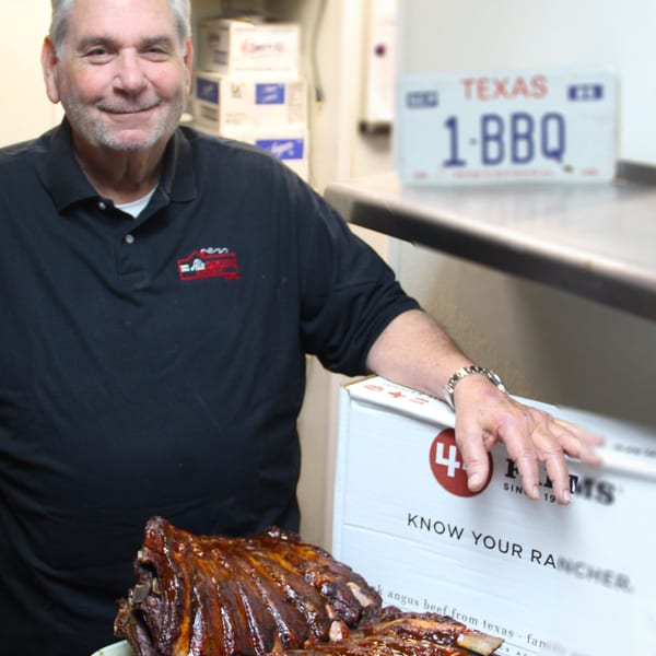 Smiling man in a black polo shirt with a plate of BBQ ribs and a "1-BBQ" Texas license plate.