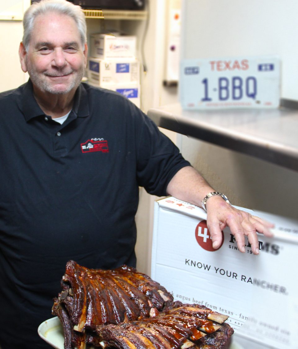 Smiling man in a black polo shirt with a plate of BBQ ribs and a "1-BBQ" Texas license plate.