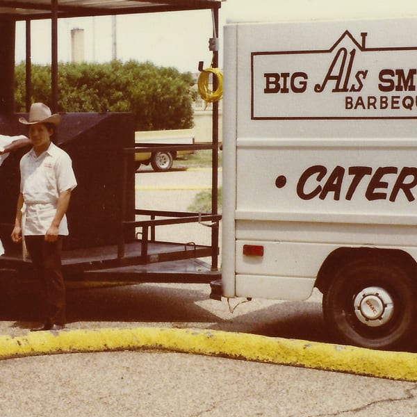 Two men in cowboy hats and aprons stand by a Big Al's Smoke House Barbeque catering truck.