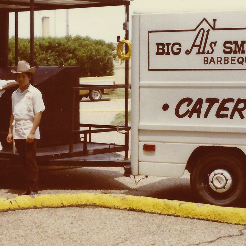 Two men in cowboy hats and aprons stand by a Big Al's Smoke House Barbeque catering truck.
