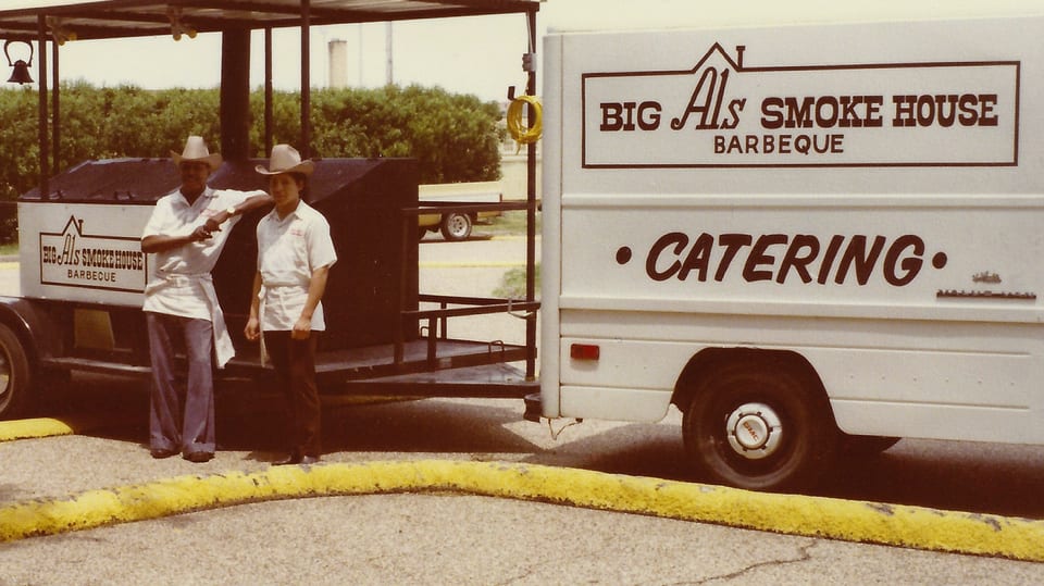Two men in cowboy hats and aprons stand by a Big Al's Smoke House Barbeque catering truck.
