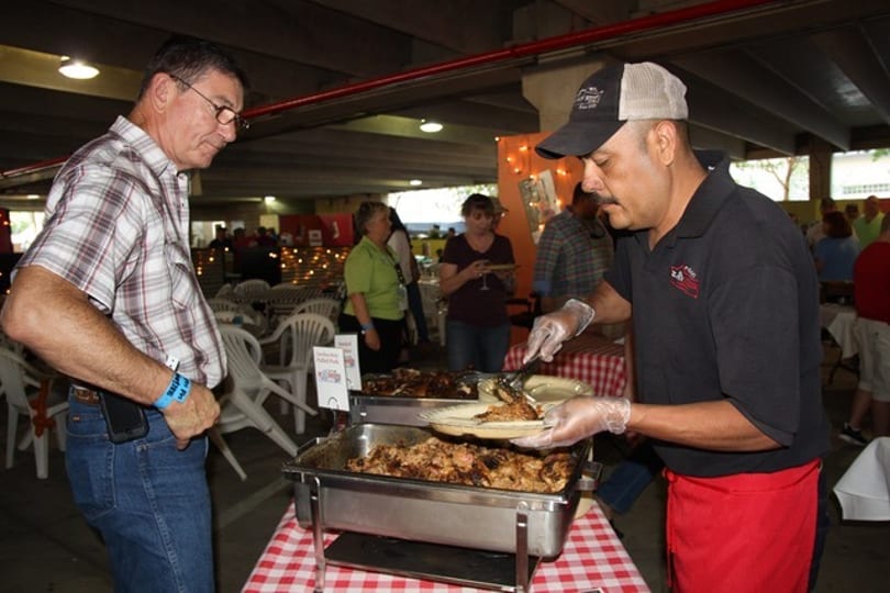 A chef serves grilled chicken from a buffet to a man in a plaid shirt at an event.