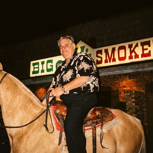 Man on a horse with a handler, in front of Big Q's Smokehouse at night.