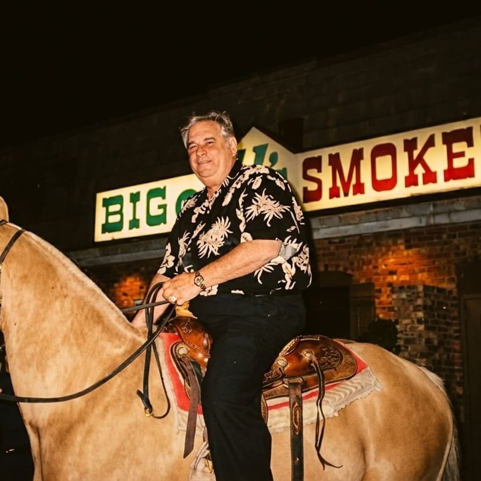Man on a horse with a handler, in front of Big Q's Smokehouse at night.