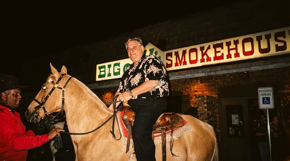 Man on a horse with a handler, in front of Big Q's Smokehouse at night.