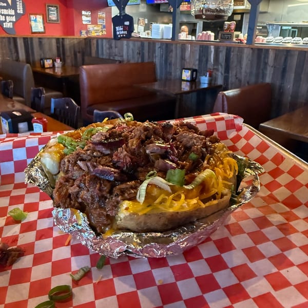 A loaded baked potato with pulled pork, cheese, and green onions on a checkered tray in a restaurant.
