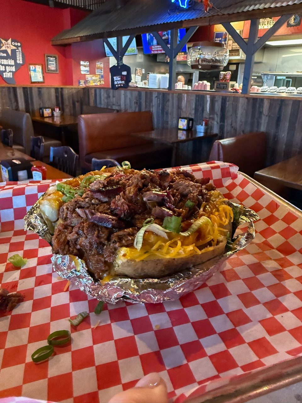 A loaded baked potato with pulled pork, cheese, and green onions on a checkered tray in a restaurant.