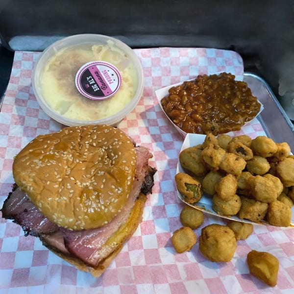 A BBQ meal with a brisket sandwich, baked beans, fried okra, and banana pudding on a checkered tray.