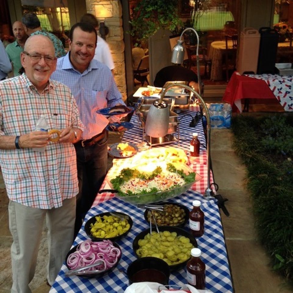 Two men smiling next to a buffet table laden with salads, condiments, and bread rolls.