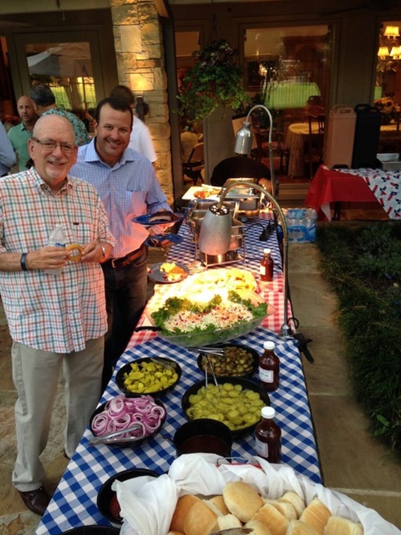 Two men smiling next to a buffet table laden with salads, condiments, and bread rolls.
