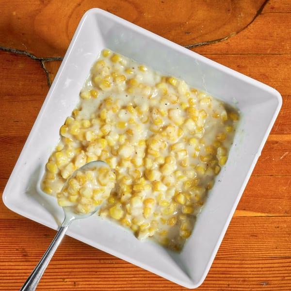 Creamed corn in a white square bowl with a spoon, on a rustic wooden table.
