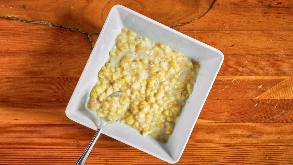 Creamed corn in a white square bowl with a spoon, on a rustic wooden table.