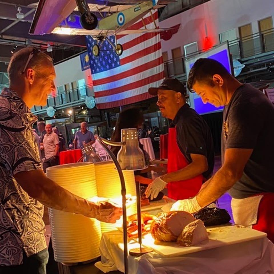 Two chefs carve meat at a buffet station with a guest, under a hanging airplane and American flag.
