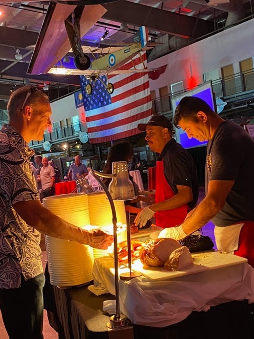 Two chefs carve meat at a buffet station with a guest, under a hanging airplane and American flag.