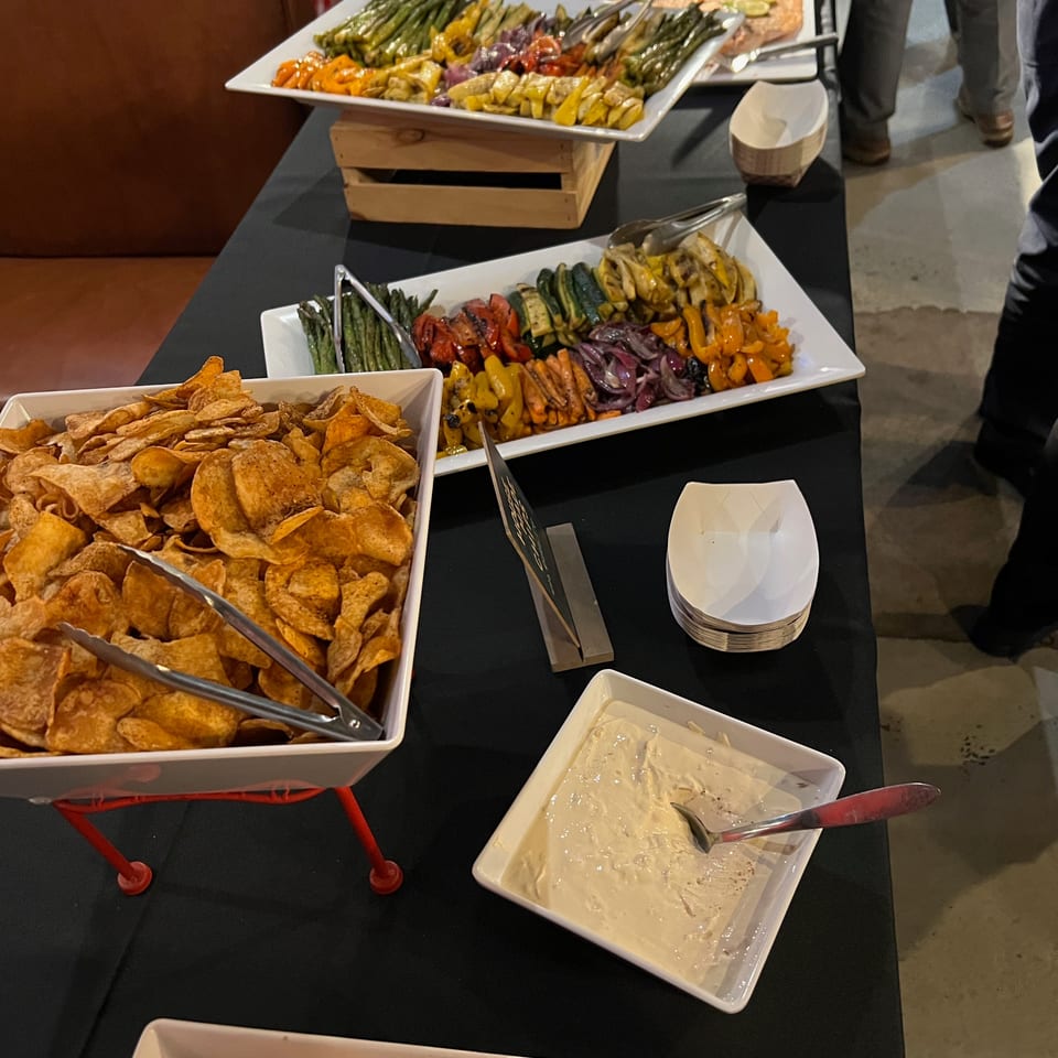 Buffet table with chips, dip, roasted vegetables, and salmon, with people in the background.