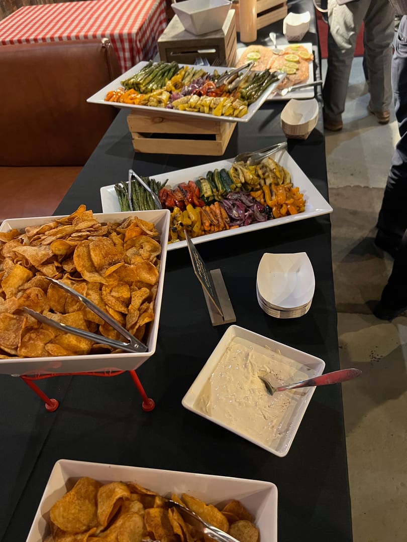Buffet table with chips, dip, roasted vegetables, and salmon, with people in the background.