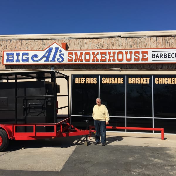Man stands beside a large red and black smoker on a trailer in front of Big Al's Smokehouse Barbecue.