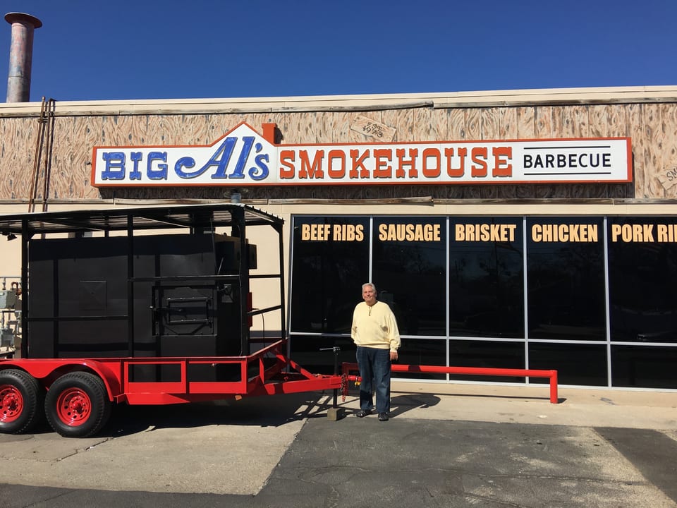 Man stands beside a large red and black smoker on a trailer in front of Big Al's Smokehouse Barbecue.