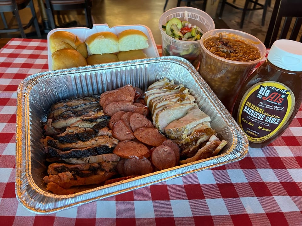 Big Al's BBQ family meal with sliced brisket, sausage, turkey, rolls, baked beans, and cucumber salad.