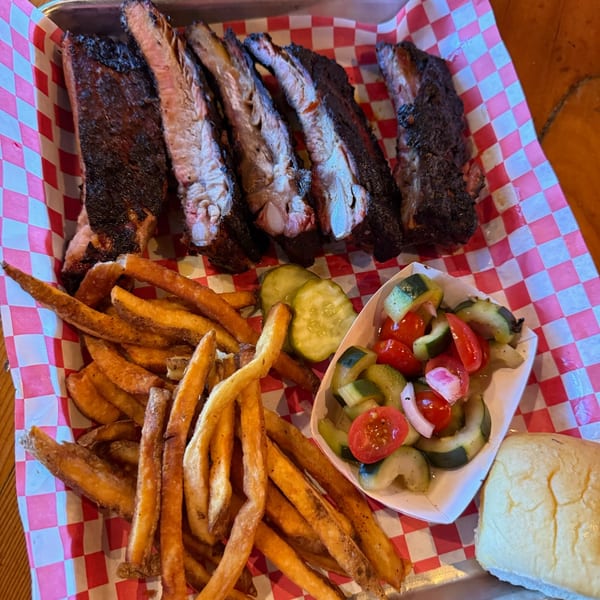 Smoked ribs, crispy fries, cucumber and tomato salad, and a dinner roll on a checkered tray.