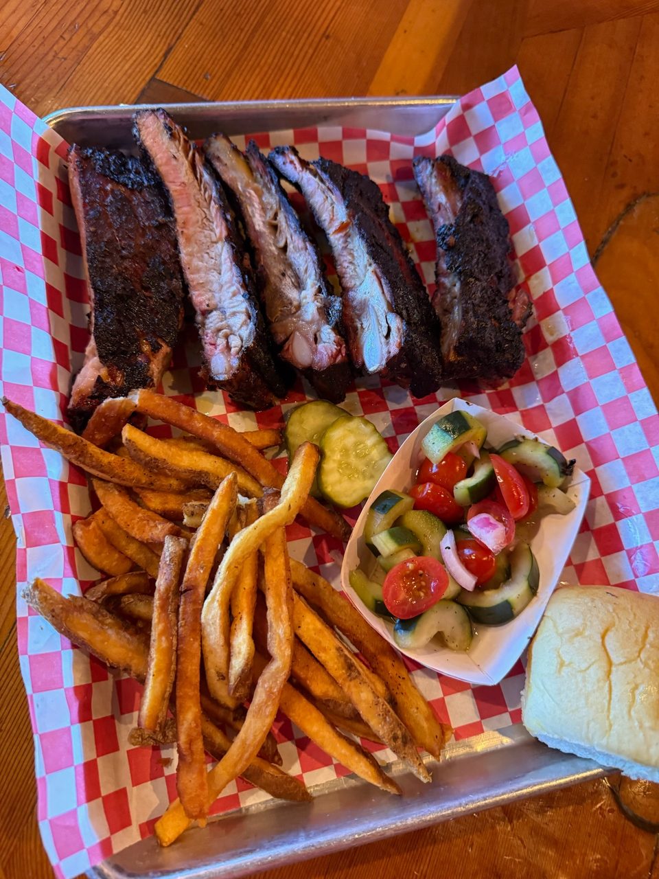 Smoked ribs, crispy fries, cucumber and tomato salad, and a dinner roll on a checkered tray.