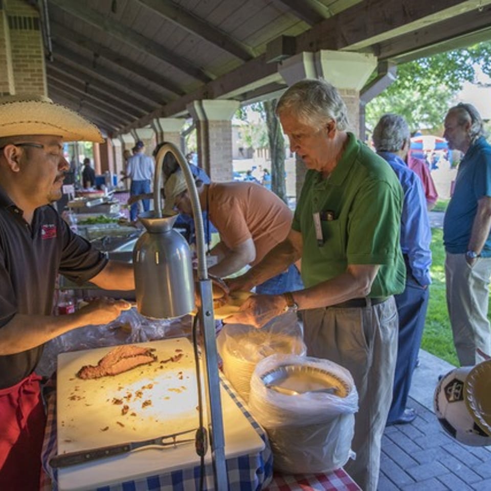 A man in a sombrero carves meat at an outdoor buffet, serving a line of people.