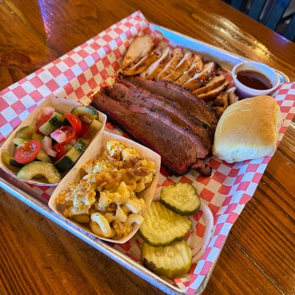 A BBQ platter with sliced brisket, chicken, mac and cheese, cucumber salad, pickles, and a roll.
