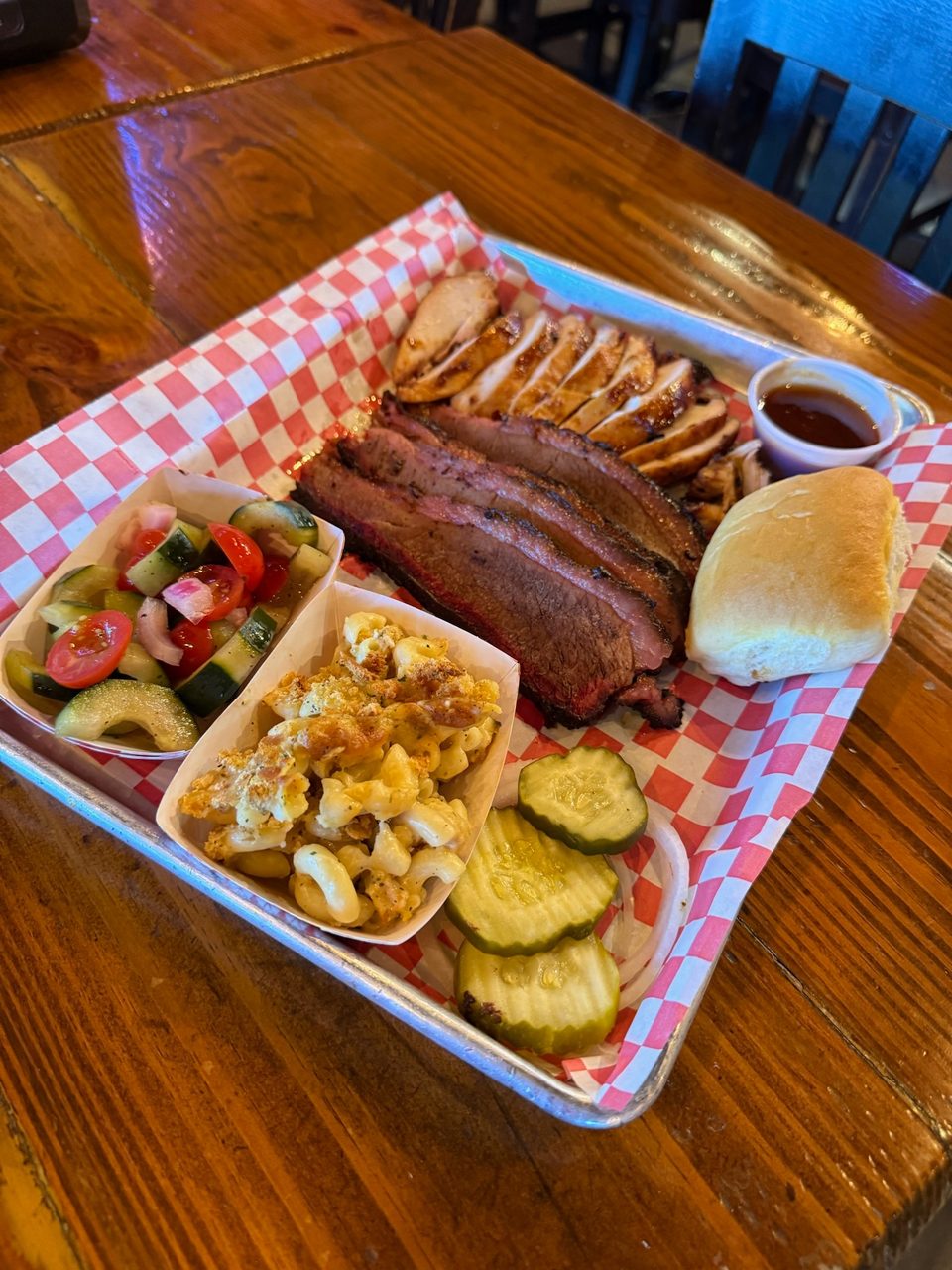 A BBQ platter with sliced brisket, chicken, mac and cheese, cucumber salad, pickles, and a roll.