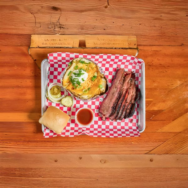 Brisket plate with a loaded baked potato, pickles, onions, a roll, and BBQ sauce on a tray.