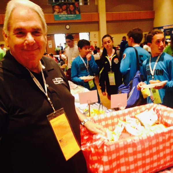 Man in black polo shirt smiling at camera, with people and a red checkered basket in background.