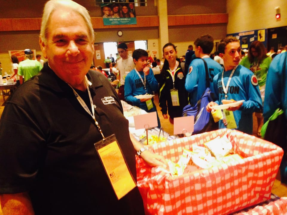 Man in black polo shirt smiling at camera, with people and a red checkered basket in background.