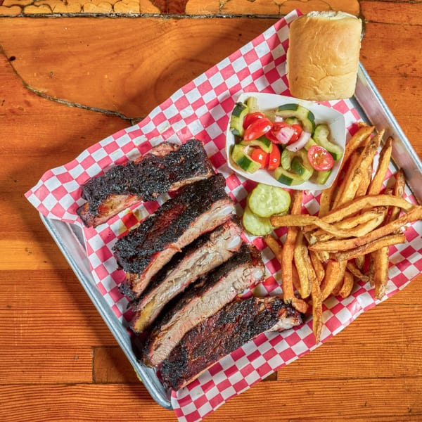 Smoked ribs, french fries, cucumber salad, and a bun on a metal tray with red and white checkered paper.