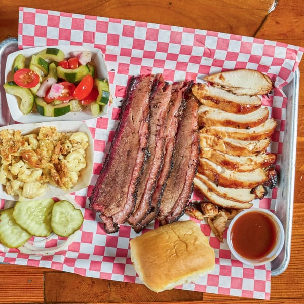 A BBQ platter with sliced brisket, chicken, mac and cheese, cucumber salad, pickles, and a roll.