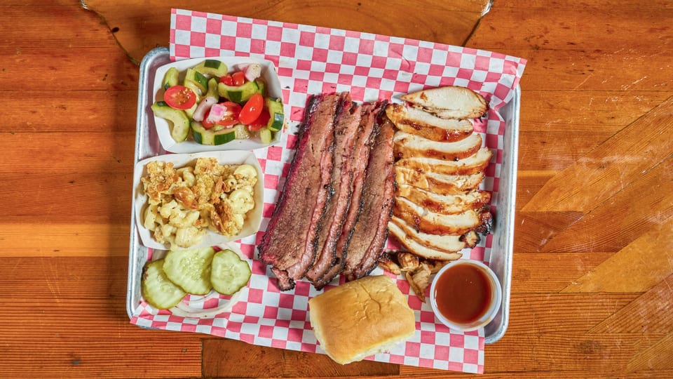 A BBQ platter with sliced brisket, chicken, mac and cheese, cucumber salad, pickles, and a roll.
