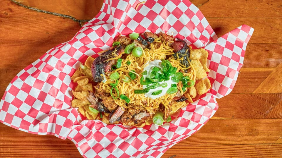 BBQ nachos with pulled pork, brisket, shredded cheese, sour cream, and green onions in a red checkered basket.