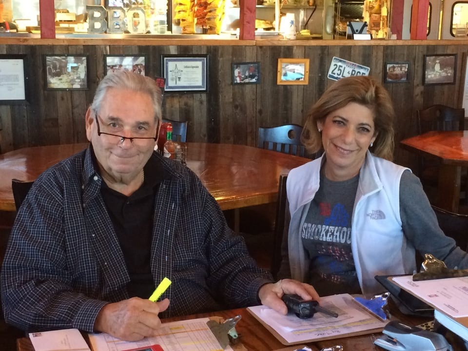 Two smiling people, a man and a woman, seated at a wooden table in a rustic restaurant.