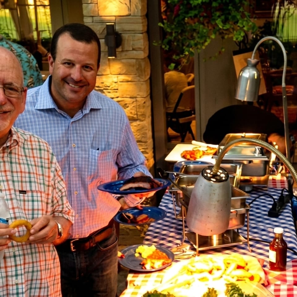 Two men smiling at an outdoor buffet with chafing dishes and food on checkered tablecloths.