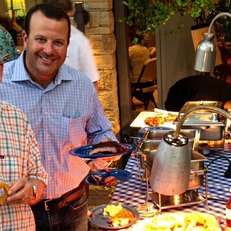 Two smiling men at a buffet table with barbecue ribs, onion rings, and other food.