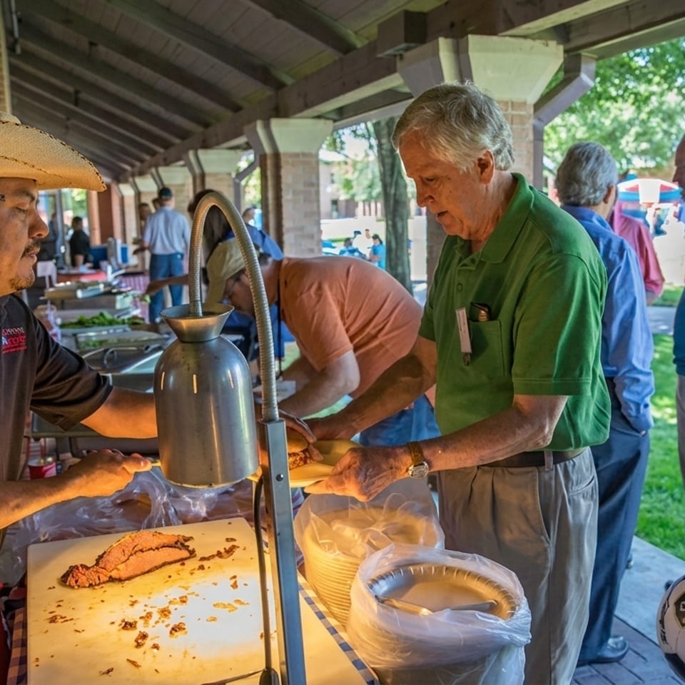 A man in a sombrero serves sliced brisket to guests at an outdoor buffet line.