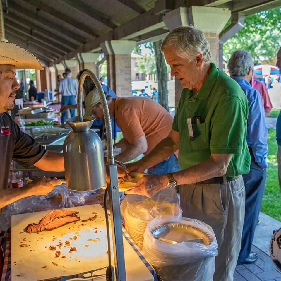 A man in a sombrero serves brisket to guests at an outdoor barbecue buffet.