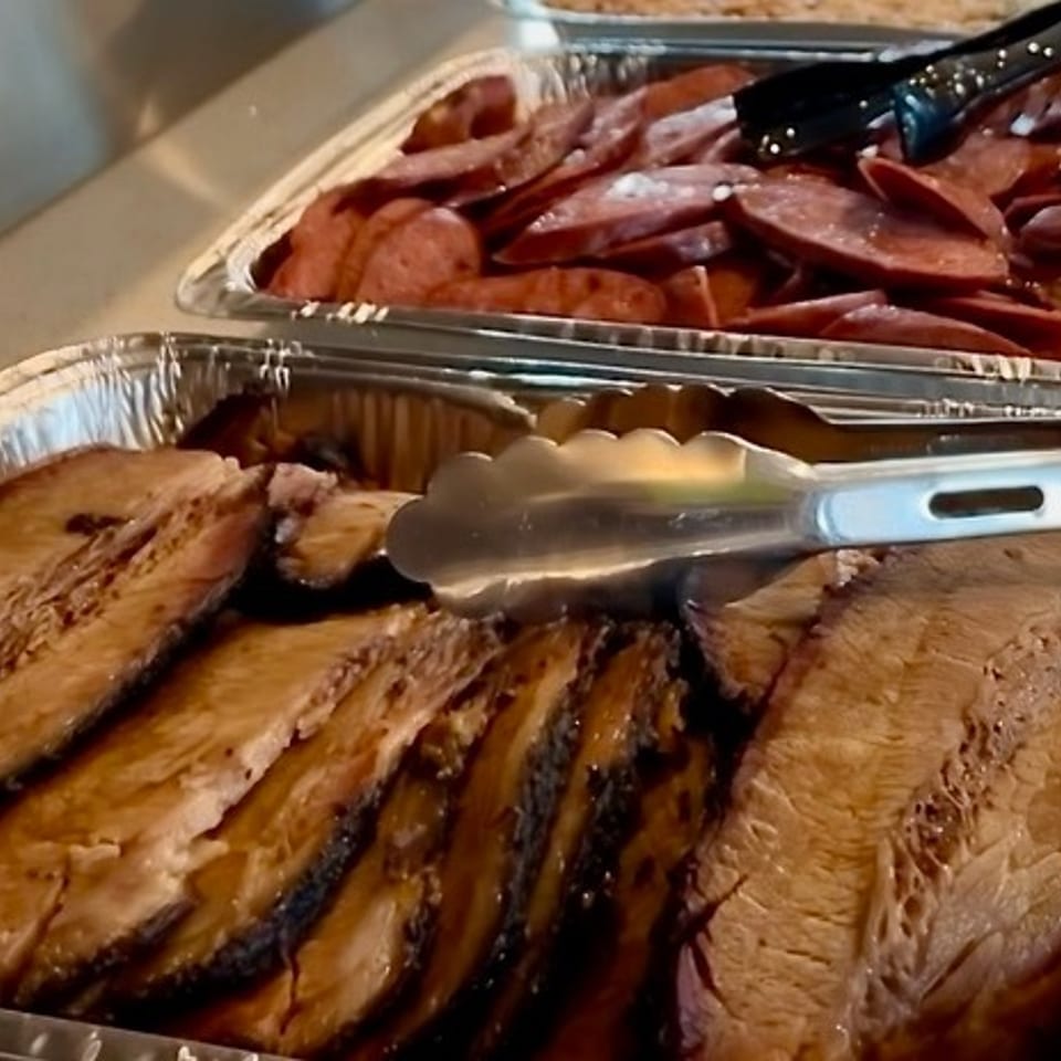 Two foil pans filled with sliced brisket and sausage, with serving tongs.