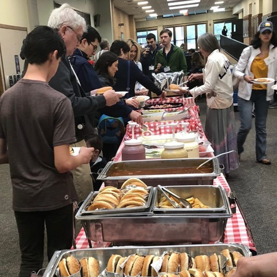 People serving themselves hot dogs and other food from a buffet line with a red and white checkered tablecloth.