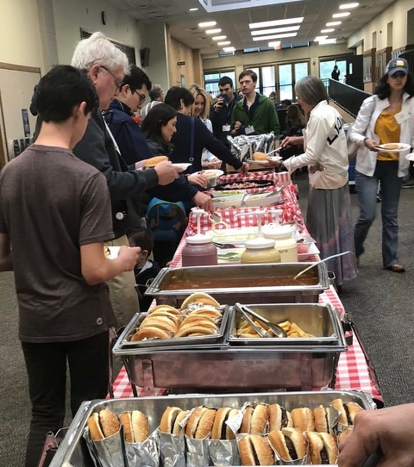 People serving themselves hot dogs and other food from a buffet line with a red and white checkered tablecloth.
